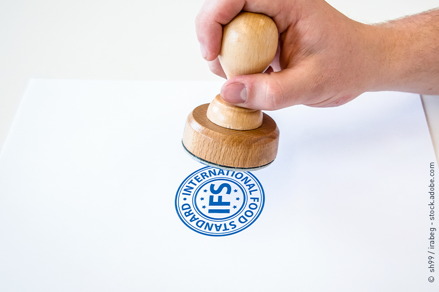 Eine Hand hält einen Stempel und stempelt das IFS-Logo auf ein weißes Blatt Papier.