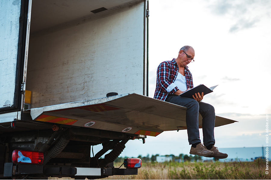 Das Bild zeigt einen LKW-Fahrer. Er sitzt entspannt auf der Ladeklappe seines LKW und liest ein Buch.
