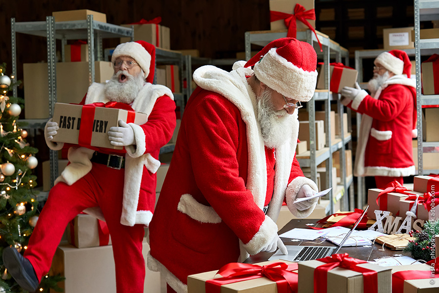 Zwei Weihnachtmänner im Lager räumen Pakete aus und ein. Ein weiterer Weihnachtsmann tippt etwas in den Laptop ein