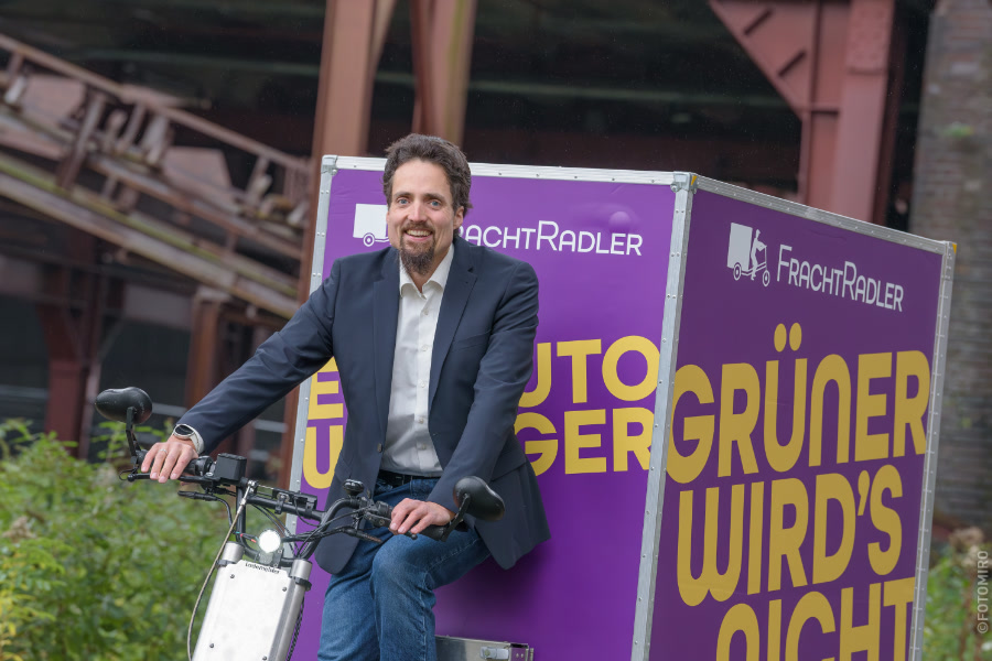 Roman Ebener auf einem Lastenrad mit einem großen lila Werbeschild von FrachtRadler.