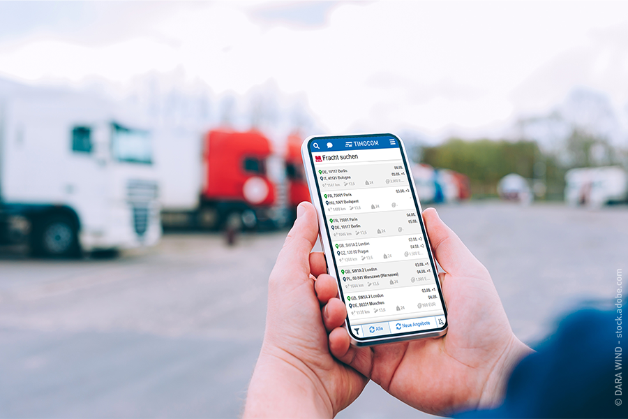 Auf einem Smartphone ist die TIMOCOM Frachtenbörse zu sehen. Dahinter stehen LKW (c) DARA WIND – stock.adobe.com / Frei zur redaktionellen Verwendung im Zusammenhang mit dieser Meldung.
